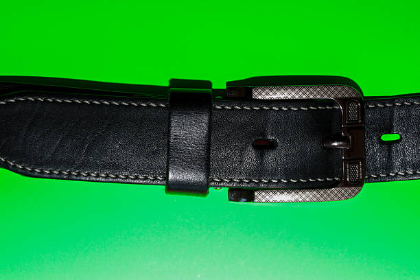 ​A classic men's belt made of black leather with contrasting white stitching and a massive metal buckle, placed on a bright green background.