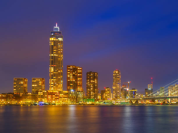 Night cityscape of Rotterdam with modern skyscrapers illuminated at dusk. Vibrant lights reflected in the Maas river. Professional architectural and urban landscape photography.