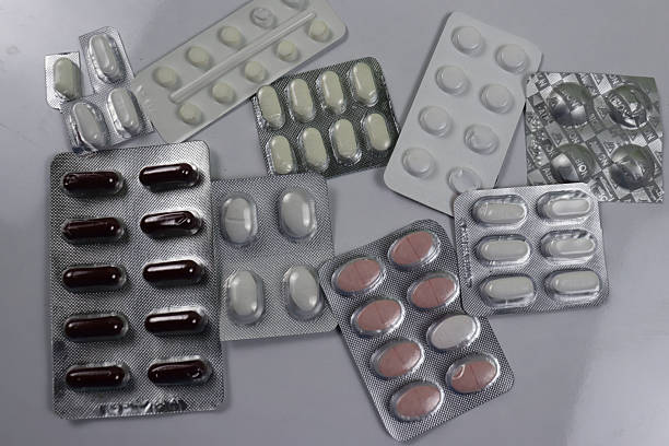 The image shows a collection of various pills and capsules in different blister packs The packs are made of silver foil and contain white pink and dark brown pills and capsules They are arranged on a plain gray surface