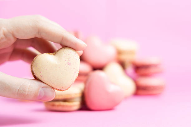 A Hand Gently Cradles a Delightful Heart-Shaped Cookie, Beautifully Set Against a Soft Pink Backdrop. Behind It, a Variety of Similar Cookies in Different Shades of Pink Create a Charming Display, Perfect for a Sweet Treat Moment Filled With Love and Joy