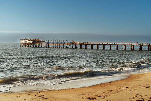Swakopmund pier stretches along a sandy beach in Namibia as gentle waves roll ashore and a few people stroll the coastline under a vast blue sky at golden hour.