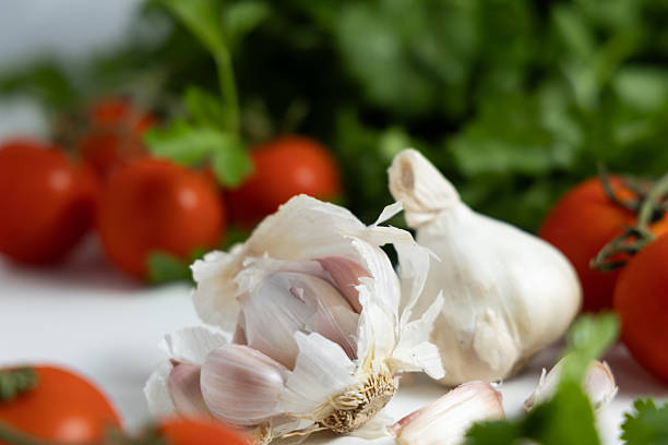 Fresh garlic cloves rest near ripe tomatoes and green herbs on a white surface. Cooking ingredients are ready for use.