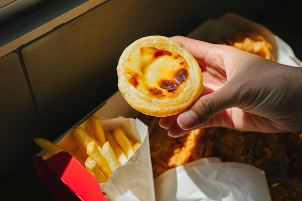 Egg tart dessert held by hand with fried chicken and french fries in paper packaging on table