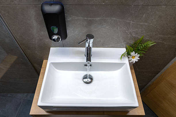 A contemporary white ceramic sink with a shiny chrome faucet is installed in a modern bathroom with gray walls, a soap dispenser, and a small decorative plant