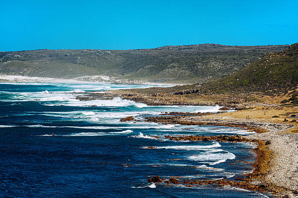 Dramatic coastal landscape at Cape Point near Cape Town,  rugged cliffs, crashing ocean waves and a rocky shoreline under a clear blue sky.