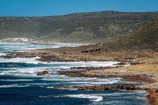 Dramatic coastal landscape at Cape Point near Cape Town,  rugged cliffs, crashing ocean waves and a rocky shoreline under a clear blue sky.