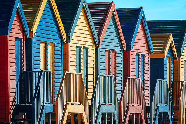 Colorful wooden beach huts lined along Muizenberg Beach in Cape Town, South Africa, showcasing vibrant painted facades, repeating gabled roofs and seaside charm in bright midday light.