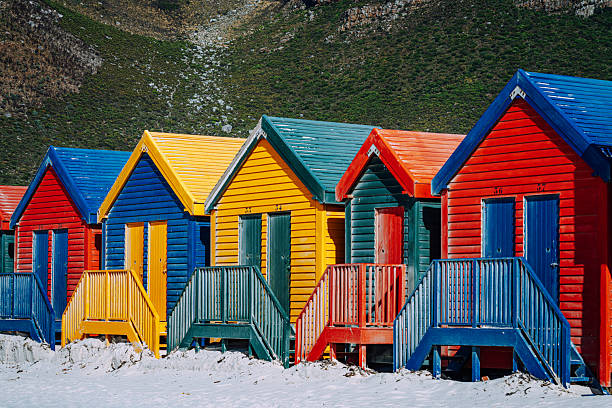 Vibrant row of colorful beach huts on Muizenberg Beach in Cape Town, South Africa, featuring painted wooden cabins and steps against a rugged hillside — a lively seaside summer and travel scene.