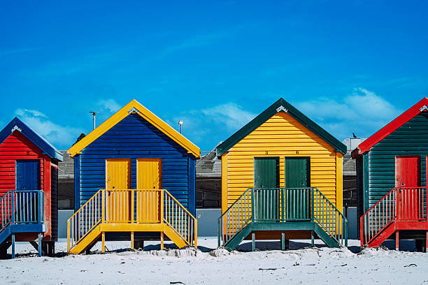 Vibrant row of painted beach huts on Muizenberg Beach in Cape Town, South Africa, contrasted against a bright blue sky and white sand, evoking summer, travel and seaside nostalgia.