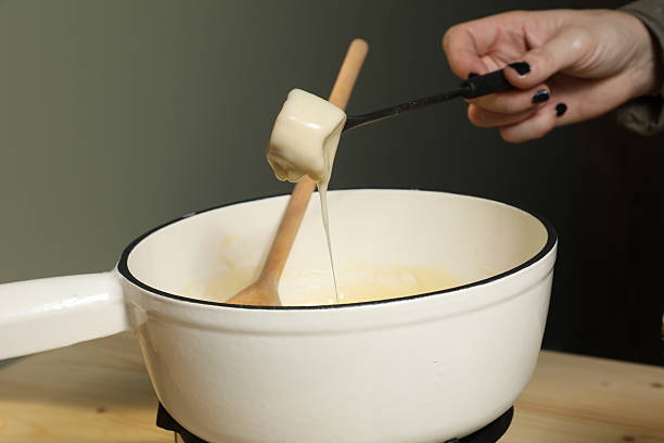 Une personne prépare une fondue bourguignonne en plongeant un morceau de fromage dans le mélange. La scène se déroule dans une cuisine avec des ustensiles simples.