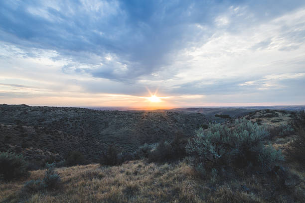 A beautiful sunset over a desert landscape. The sky is filled with clouds, and the sun is setting in the distance. Scene is peaceful and serene, as the sun's rays cast a warm glow over the landscape