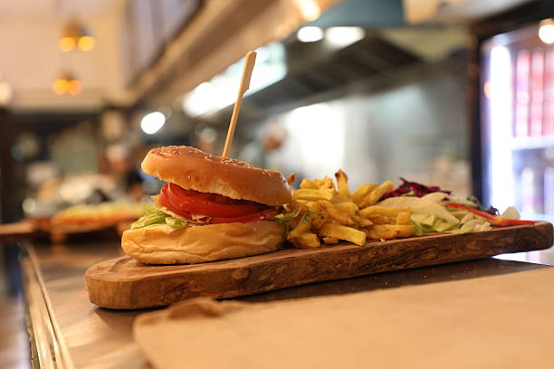 Un hamburger avec de la laitue et des tomates est placé sur une planche en bois. À côté, des frites sont disposées. L'environnement montre un lieu de restauration rapide en arrière-plan.