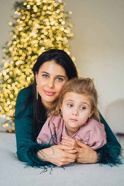 A woman and her daughter sit close together on a soft surface. They look at the camera while a Christmas tree with bright lights fills the background.