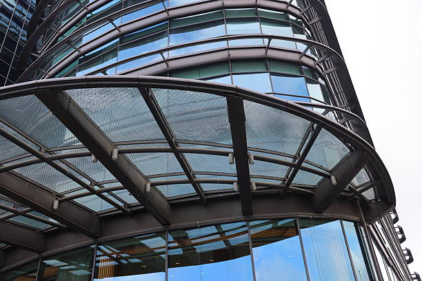 the architectural detail showcases the intersection of contemporary glass and steel structures under daylight. the composition features a high-rise building with a dark-tinted glass curtain wall reflecting the surrounding environment. in the foreground, a large geometric canopy constructed from a metal frame and perforated metal sheets provides a structural overhead element. curved architectural lines of an adjacent building section create a sense of fluid movement against the rigid grid of the skyscraper windows. the use of industrial materials like aluminum and tempered glass highlights a clean, urban aesthetic typical of modern commercial or office districts.