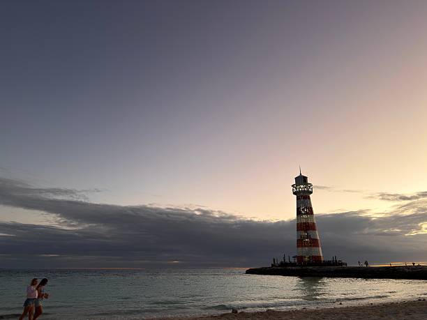 Ocean Cay, Bahamas - January 8, 2026: Two young adult women enjoying a relaxing evening walk on the sandy beach as the iconic red and white lighthouse illuminates the beautiful tropical sunset