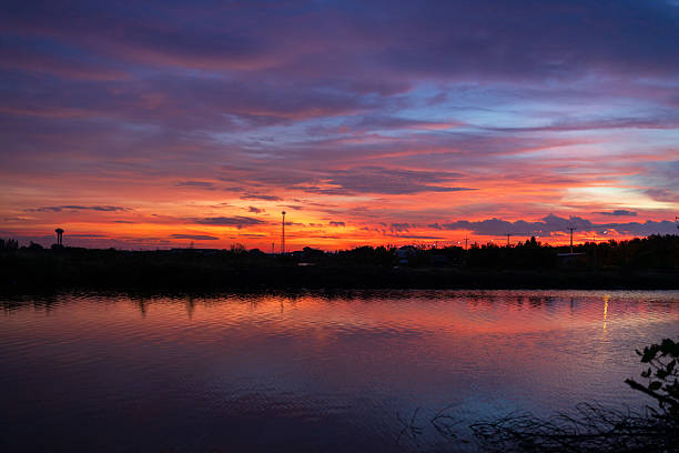 A breathtaking sunset scene featuring a dramatic sky filled with orange, purple, and blue hues, perfectly reflected on the tranquil surface of a lake or pond with a silhouette of a suburban horizon.