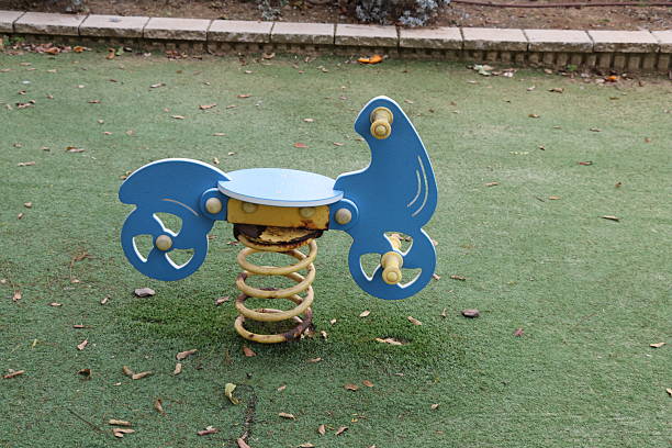 Blue spring rider playground toy designed to resemble a motorcycle. Made of durable plastic, it sits on a coiled metal spring, allowing for bouncing movement. Situated on a lush, grassy area with scattered leaves. A stone border separates the grass from a garden bed.