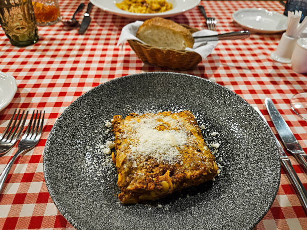 Traditional lasagna with meat sauce and grated parmesan served on a dark plate over a red checkered tablecloth in an Italian restaurant. Rustic dining atmosphere with bread and table setting.