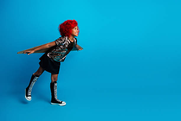 A lively young girl with bright red hair wears a sparkling top and black skirt, balancing on a board against a vivid blue backdrop. This playful image conveys energy joy and fashion flair.
