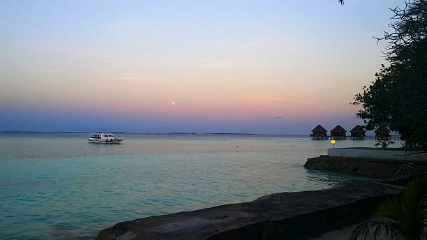 Tropical sunset over calm ocean with small boat and overwater villas in distance, peaceful luxury travel destination.