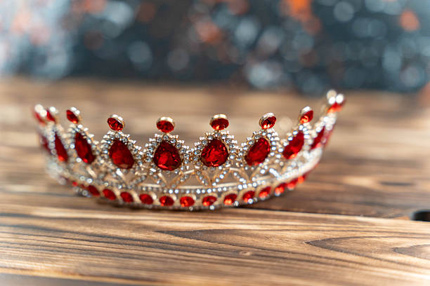 Silver tiara featuring red gemstones displayed on a wooden surface, highlighting its intricate design and sparkling details with a blurred colorful backdrop