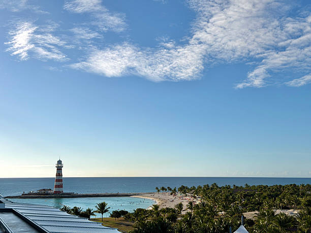 Ocean Cay lighthouse standing on a pier, overlooking a white sand beach filled with sunbathers, palm trees, and turquoise blue ocean water under a clear sky