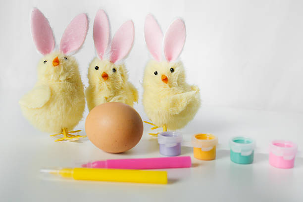 One Easter egg with bright colorful paints, markers and funny small toy chicks with Easter bunny ears on white background