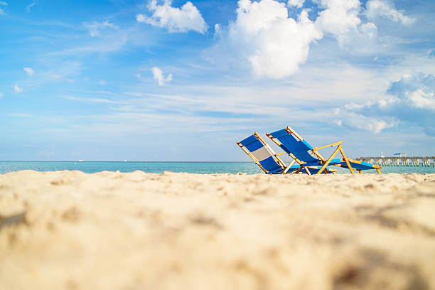 Two blue beach chairs on a Florida white sand beach near turquoise ocean water and a long wooden pier under a clear blue sky. Bright morning sun highlights the tropical coastline, offering a perfect holiday atmosphere for summer travel marketing.
