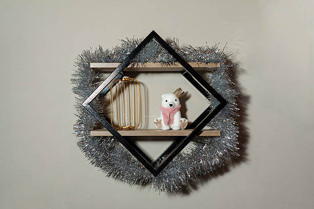 Festive wall shelf with silver tinsel wreath, geometric frame, small polar bear toy and gold lantern on a neutral wall interior background.