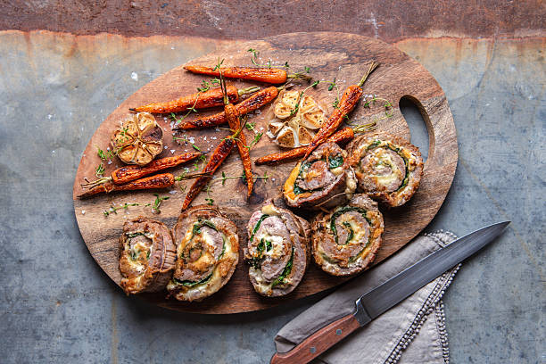 Stuffed Pork Roulade Slices With Roasted Baby Carrots and Garlic on Wooden Cutting Board