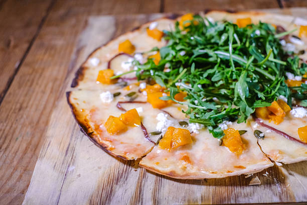 Crispy flatbread pizza topped with fresh arugula, roasted butternut squash, melted cheese and goat cheese crumbles on a rustic wooden board. Warm, savory flavors are highlighted by a thin, charred crust and scattered pepitas, making an appetizing, seasonal dish ideal for casual dining and food photography.