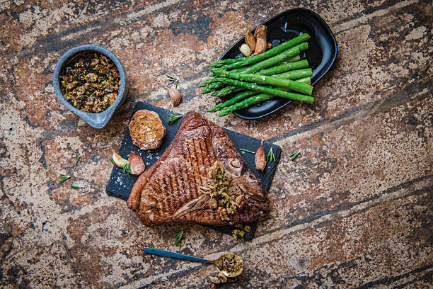 Grilled T-bone steak plated on a slate board with roasted garlic, chimichurri sauce and steamed asparagus over a textured rustic surface. Top-down food photography that conveys hearty, gourmet dinner vibes, savory flavors and a restaurant-style presentation ideal for menus, blogs and recipe content.