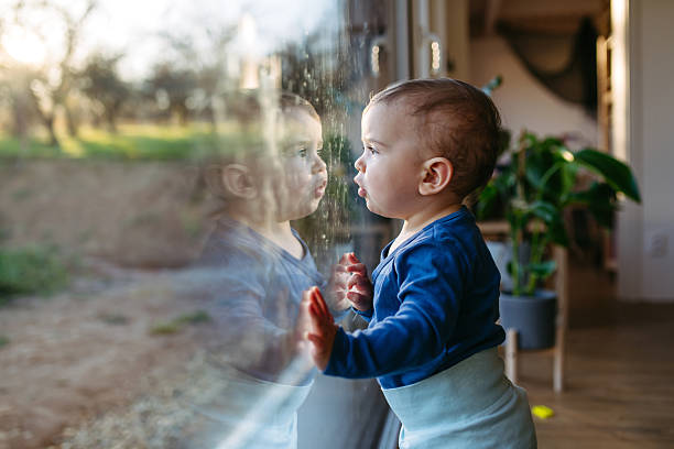 A young child in a sleeveless striped shirt touching a glass surface, with their reflection visible.
