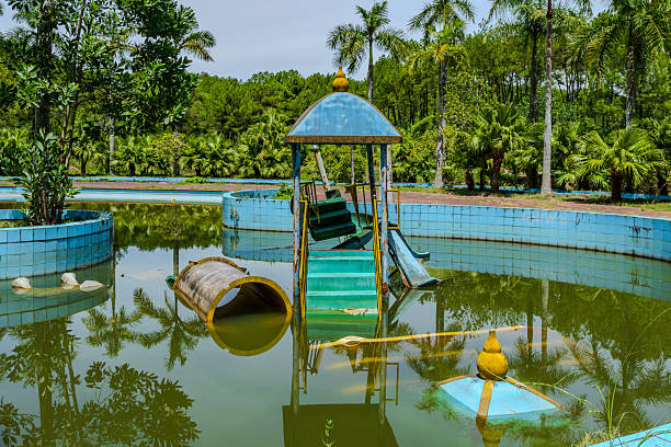 Moody view of an abandoned water park in Vietnam, featuring decaying structures, overgrown vegetation and a mysterious atmosphere. The empty pathways, weathered slides and tranquil lake create a haunting yet captivating scene. Ideal for themes related to urban exploration, decay, adventure travel and forgotten places.