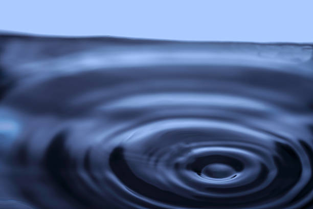 275.Close-up of water surface showing concentric ripples from droplet impact. Dark reflective water contrasts with light blue background, highlighting wave patterns and fluid motion