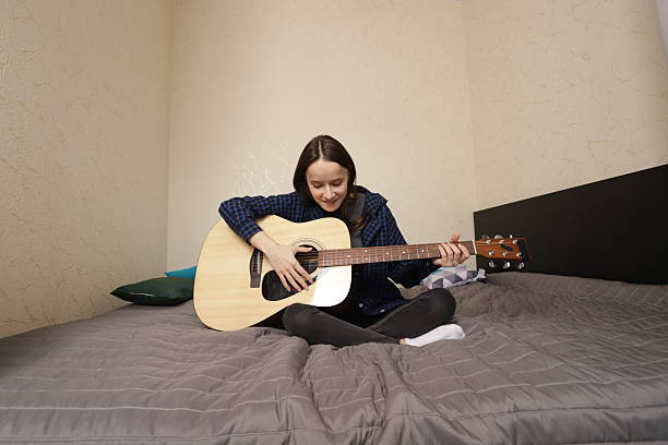focused young woman practices guitar chords on a bed, hands on strings and fretboard, learning music step by step, quiet indoor routine, self study practice in a cozy bedroom