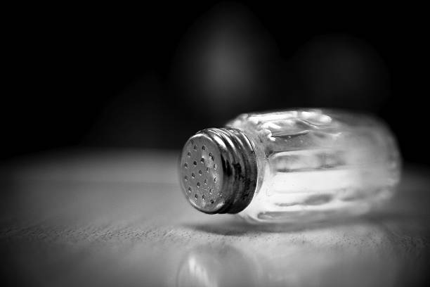 Black-and-white close-up of a glass salt shaker lying on its side on a reflective surface, shallow depth of field, soft bokeh background