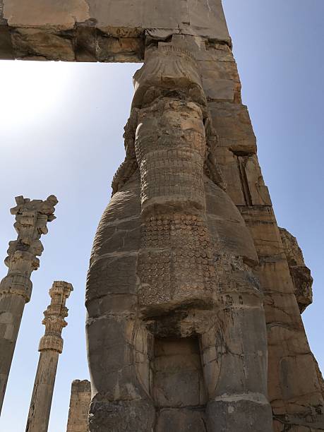 Ancient colossal stone guardian statue at gate with carved human-animal hybrid features, weathered surface, towering columns and clear blue sky