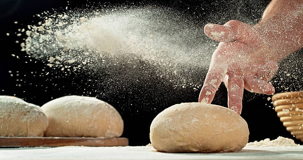 Close up of baker’s hand tossing flour over prepared risen dough on wooden table. Cinematic baking concept, artisan bread preparation and texture detail.
