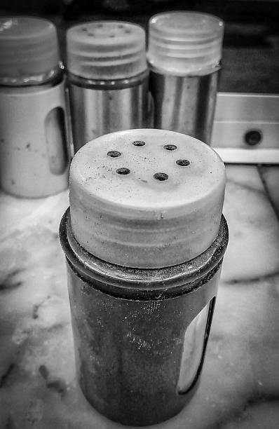 Black and white close-up of a metal and plastic shaker on a marble surface with three similar shakers blurred in background