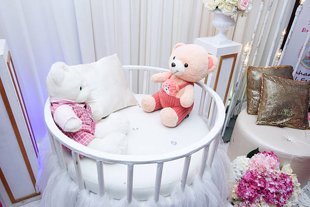 Two cute plush teddy bears sitting inside a decorated white round baby crib, surrounded by floral accents and gold pillows.