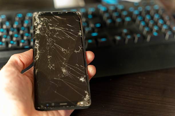 Hand displays a damaged smartphone featuring a cracked screen, positioned in front of a computer keyboard with illuminated blue keys on a dark surface