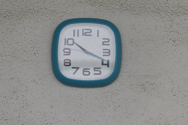 A blue square clock with the hands at 3:55. The clock is on a wall. The clock is showing the time as 3:55