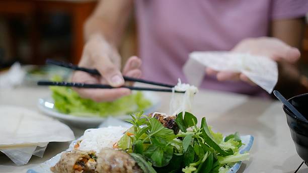 Woman enjoying fresh vietnamese spring rolls with vermicelli noodles and various herbs at a restaurant, using chopsticks to prepare a delicious healthy meal