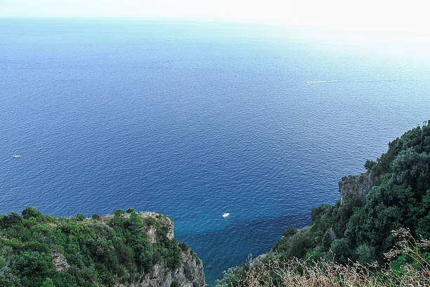 High view over calm blue ocean framed by lush rocky cliffs with small boat and large copy space. Summer travel seascape, nature background.