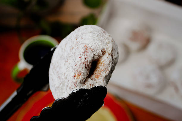 A powdered pastry filled with jelly or jam held up with tongs with coffee in the background. Paczki polish doughnuts.