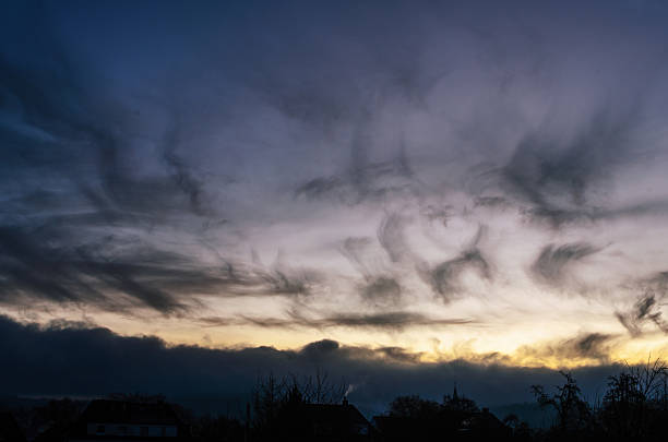 The sky displays a captivating mix of dark, wispy clouds and a soft, warm light peeking through the horizon, creating a moody atmosphere over silhouetted rooftops