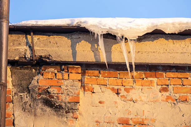 Icicles form on the edge of a building. The bricks show signs of wear. The sun shines, and the sky is clear. Snow rests on the roof in the cold weather.