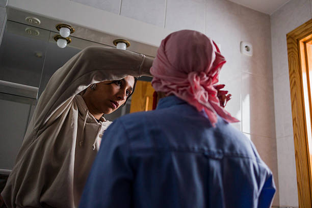 Young adult woman assisting her mature adult mother, wearing a headscarf due to cancer, in a bathroom, applying makeup, offering support and care