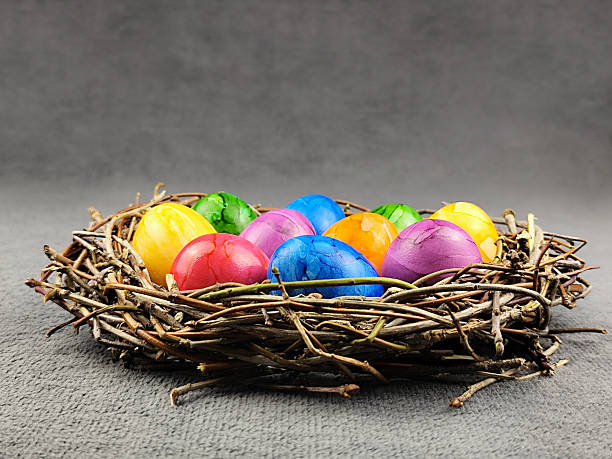 Eye-level side view of a handmade wicker nest holding a collection of colorful Easter eggs on a grey surface, highlighting the texture of the twigs.